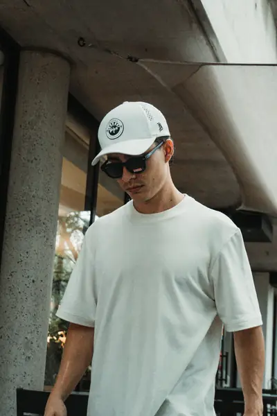 Con gorra y lentes de sol, polera blanca UEBOS bajo un techo de concreto